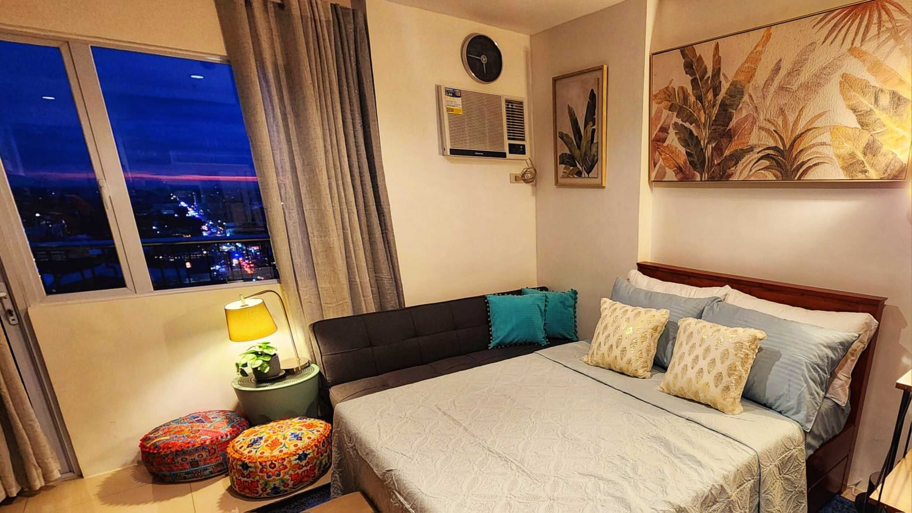 A bedroom with a large bed, a nightstand, a lamp, a window with a city view, and a wall-mounted air conditioner.