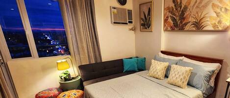 A bedroom with a large bed, a nightstand, a lamp, a window with a city view, and a wall-mounted air conditioner.
