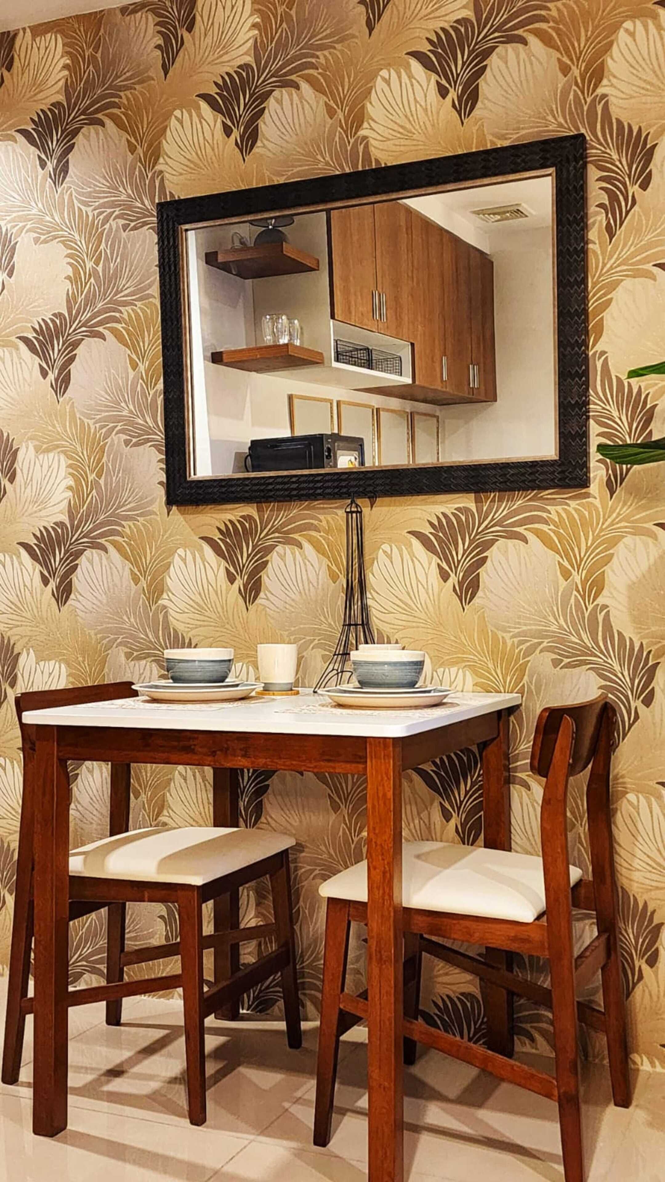 A dining area with a wooden table and chairs, a black-framed mirror above the table, and a patterned wallpaper background.
