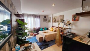 A well-lit hotel room with a bed, a desk, a chair, a large potted plant, and a window with curtains. - Bella Suite Bacolod (Bacolod)
