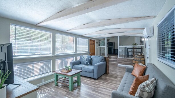 A spacious living area with a sofa, a coffee table, and a television. There are large windows with blinds, a kitchen area in the background, and a ceiling with exposed beams. - Serenity Hideaway, Charming 2bd/1ba cabin with WiFi, AC, close to shops (Ruidoso)