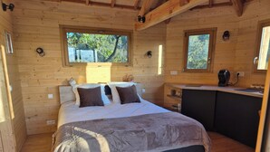 Un dormitorio de cabaña de madera con una cama, dos ventanas, un escritorio empotrado y una pequeña kitchenette.