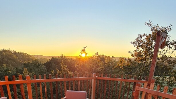 Una terraza de madera con mobiliario exterior, una mesa dispuesta con cruasanes, café y jugo, y vistas al atardecer.