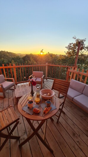 A wooden deck with outdoor furniture, a table set with croissants, coffee, and juice, and a sunset view.
