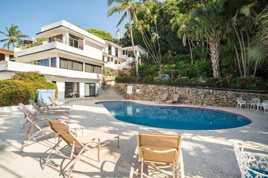 Un edificio moderno de dos pisos con piscina, rodeado de exuberante vegetación y palmeras.