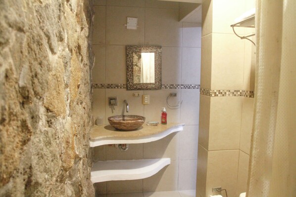 A bathroom with a stone wall, a sink with a decorative bowl, a mirror, and a shower curtain. - Villas Luna Marina (Acapulco)