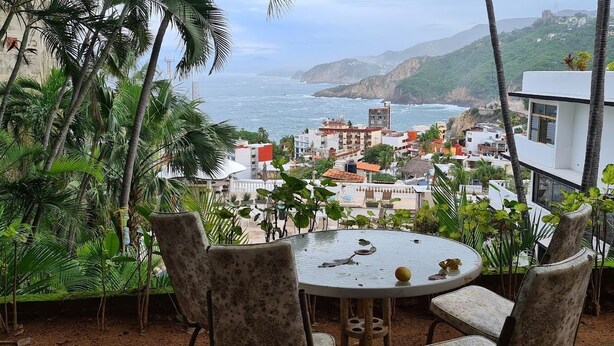 A terrace with a table and chairs overlooking a coastal town with buildings and greenery. - Villas Luna Marina (Acapulco)