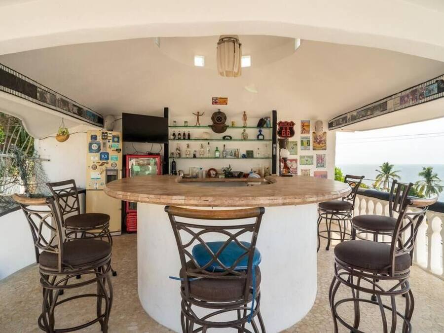 A bar area with a round table and four wicker chairs, shelves with bottles and decorations, and a view of the sea.