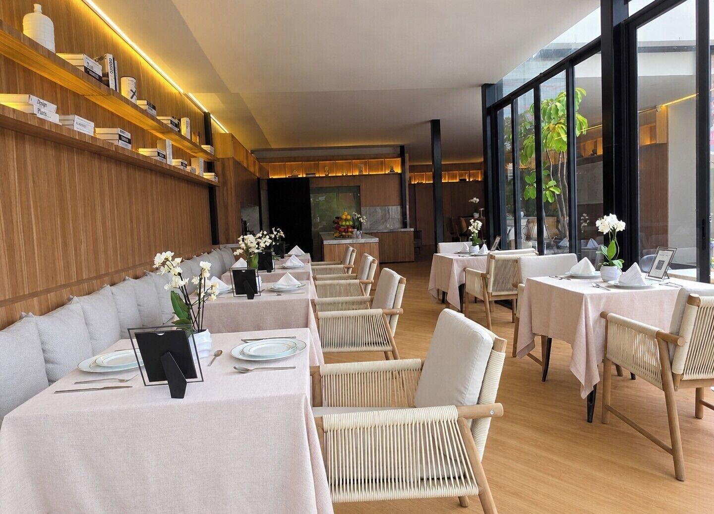Un restaurante con estanterías de madera, mesas blancas, sillas de mimbre y vistas amplias a través de grandes ventanales.