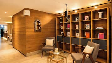 A hotel lobby with a wooden reception desk, two armchairs, a glass coffee table, and a bookshelf filled with books.