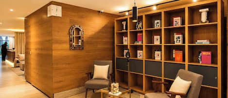 A hotel lobby with a wooden reception desk, two armchairs, a glass coffee table, and a bookshelf filled with books.