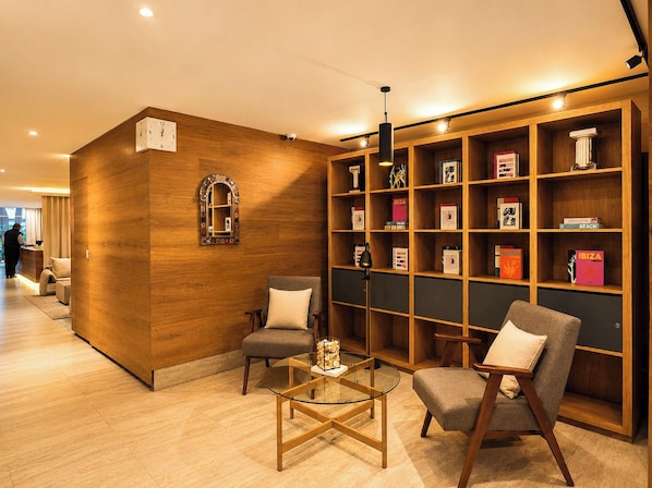 Un vestíbulo de hotel con un mostrador de recepción de madera, dos sillones, una mesa de centro de vidrio y una estantería llena de libros.