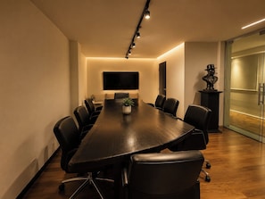 Un salón de conferencias con una mesa larga, sillas negras, una planta en maceta y un televisor de pantalla plana montado en la pared.