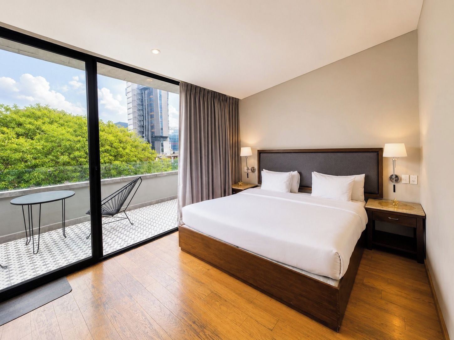 Une chambre d’hôtel moderne avec un grand lit, une vue sur la verdure et les bâtiments, et un balcon avec une chaise.