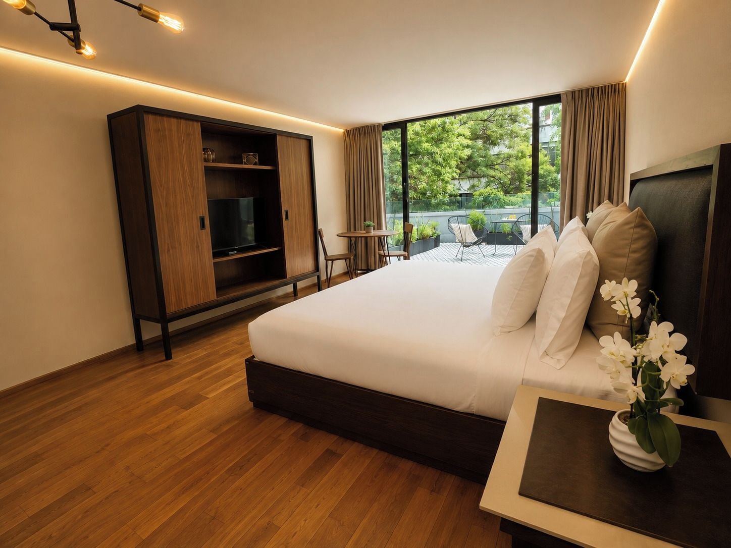 A modern hotel room with a large bed, a TV, a wardrobe, and a balcony with outdoor seating.