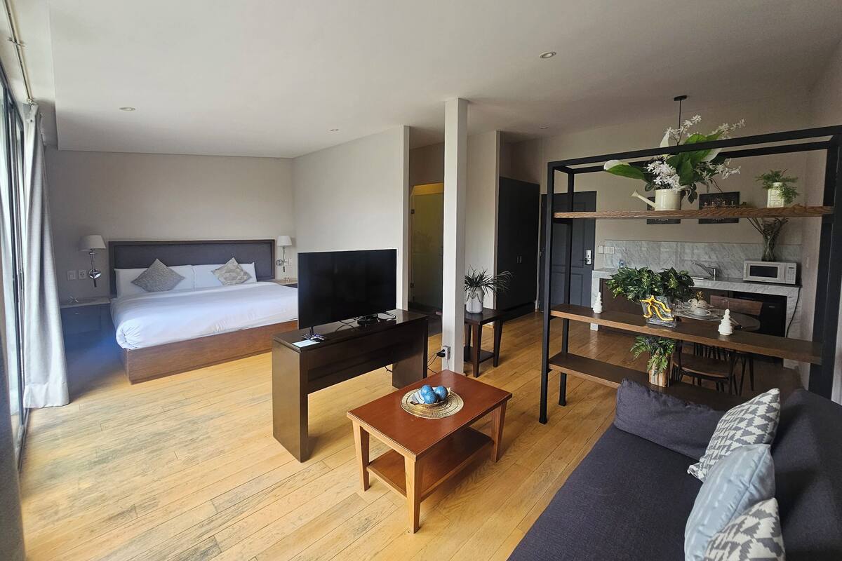 A modern hotel room with a bed, TV, wooden coffee table, sofa, and a kitchen area with a microwave.