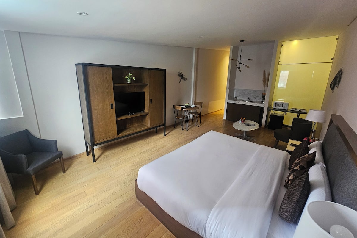 A hotel room with a large bed, a desk with a chair, a small table, a TV, and a kitchenette.