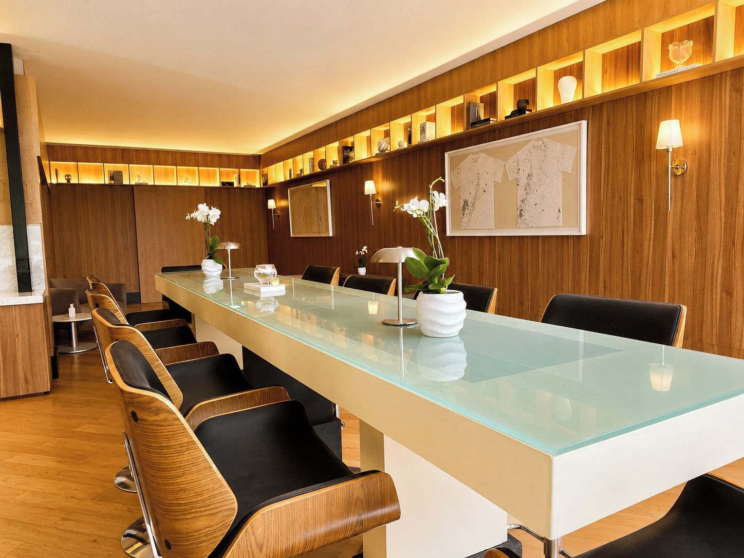 Une salle de conférence moderne dotée d’une longue table en verre, de chaises en bois et d’un mur en panneaux de bois avec des étagères.