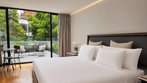 Habitación de hotel moderna con una cama grande, un balcón con mesa y sillas, y vistas a zonas verdes y edificios.
