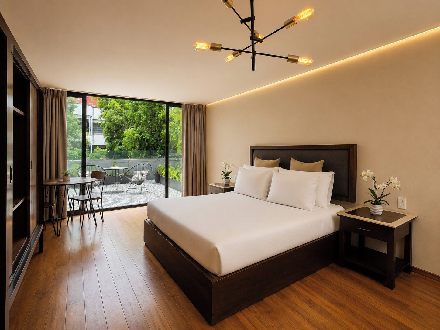 A modern hotel room with a large bed, a dining area with a table and chairs, and a view of greenery outside.