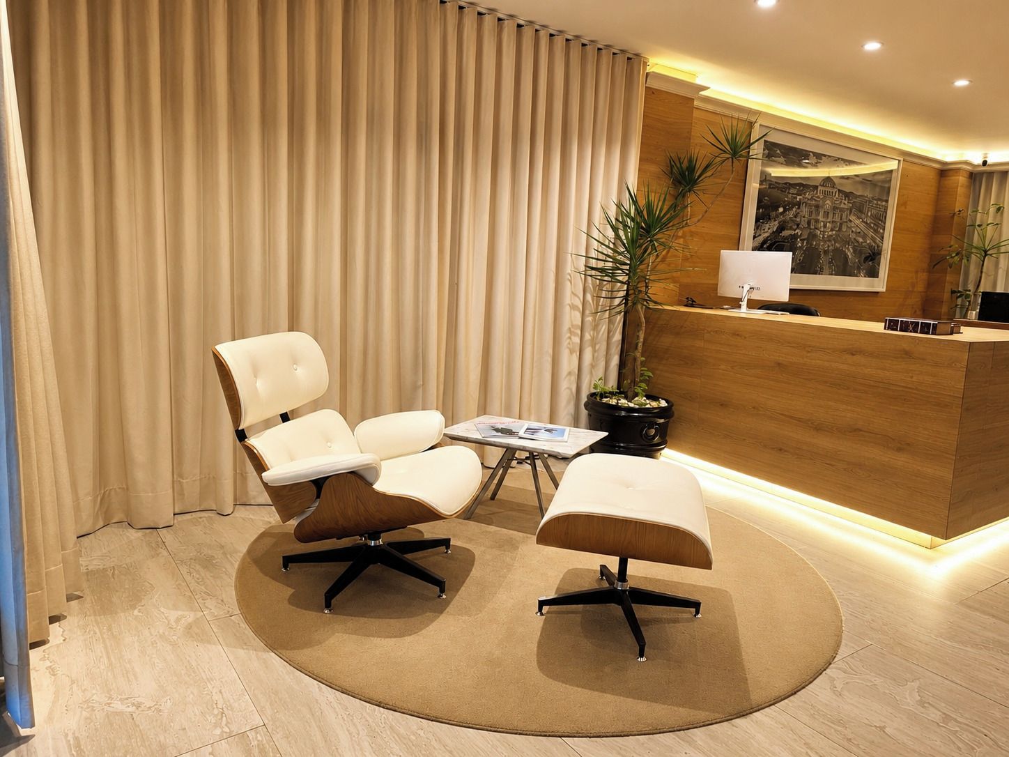 A hotel reception area with a desk, two chairs, a small table, and a potted plant.