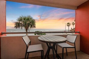 A balcony with a round wooden table and two chairs, overlooking a sunset beach with palm trees. - Beachfront Condo | Pool + Hot Tub | View (Galveston)