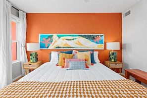 Een hotelkamer met een groot bed, oranje wanden, een kleurrijk schilderij boven het bed en twee nachtkastjes met lampen.