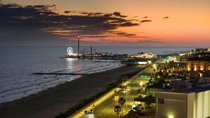 A coastal cityscape at dusk with a prominent amusement park on a pier, a busy road, and beachfront buildings. - Beachfront Condo | Pool + Hot Tub | View (Galveston)
