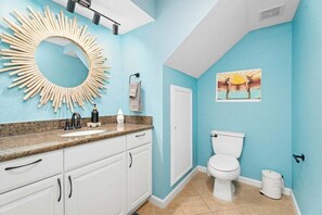 A bathroom with a blue wall, a white toilet, a sink with a granite countertop, a mirror with a sunburst design, and a picture of a sunset. - Beachfront Condo | Pool + Hot Tub | View (Galveston)