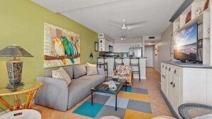 A living room with a sofa, a coffee table, and a TV. There is a kitchen area with cabinets and a microwave. - Beachfront Condo | Pool + Hot Tub | Near Seawall (Galveston)