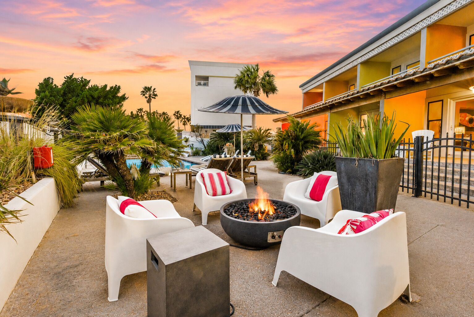 An outdoor seating area with white chairs and a fire pit, surrounded by palm trees and a pool.