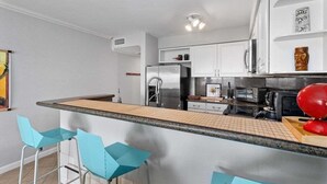 Une cuisine moderne avec des meubles blancs, un réfrigérateur en acier inoxydable, un four à micro-ondes et une machine à café. On y trouve trois tabourets de bar bleus et un comptoir en bois.