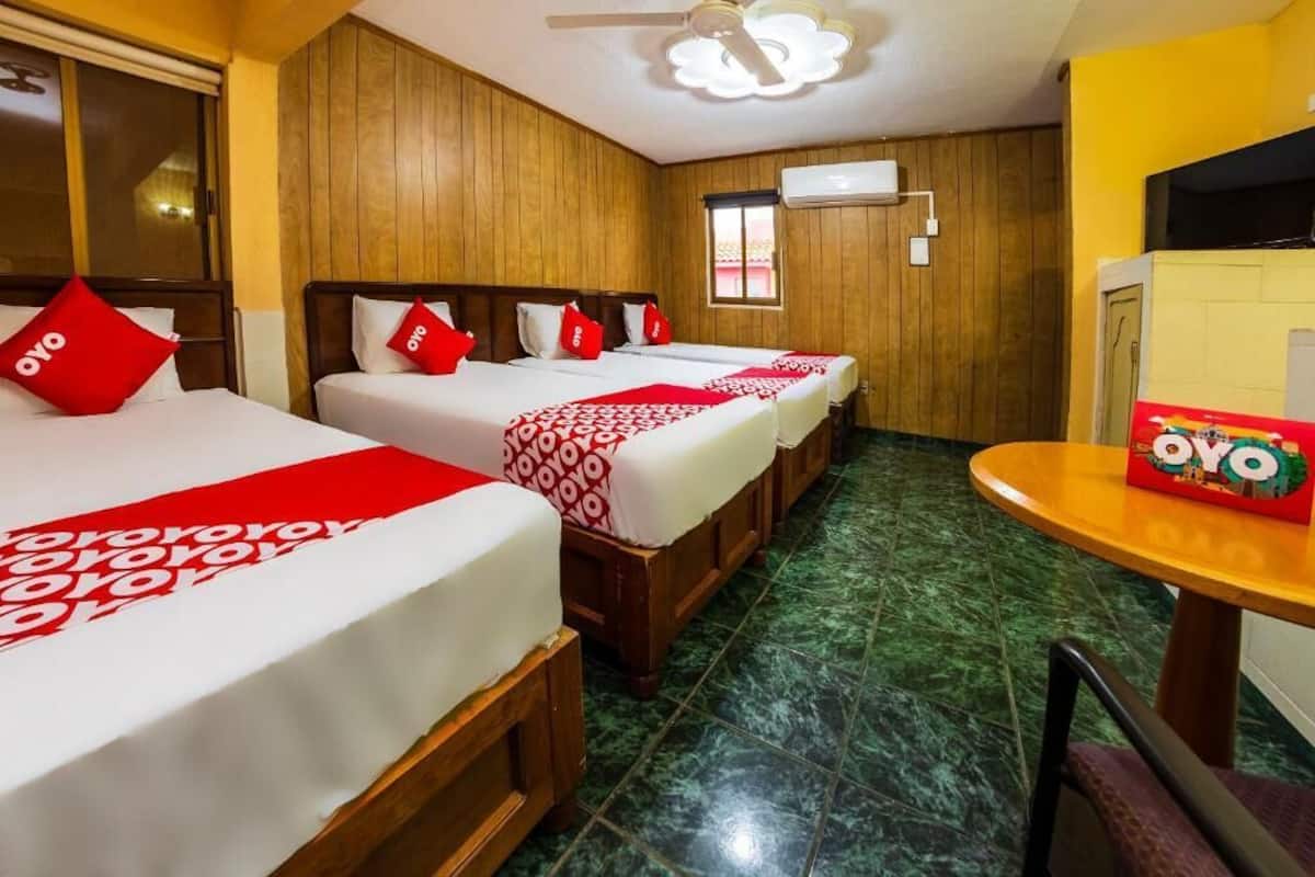 A hotel room with three beds, each with red and white bedding and matching pillows. There is a small table with a box labeled "OYO" on it.