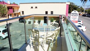Un balcon fermé par des baies vitrées, avec une table ronde et des chaises, donnant sur une rue et un immeuble portant l’enseigne « HÔTEL SAN MARCOS FAMILIAR ».