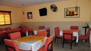 Un espace repas avec plusieurs tables et chaises, un téléviseur fixé au mur et une nature morte représentant des fruits.