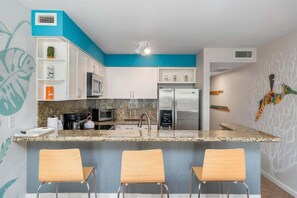 Una cocina compacta con gabinetes blancos, encimera de granito, refrigerador de acero inoxidable, microondas y cafetera. Hay tres taburetes de madera en la barra.