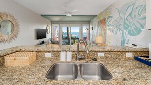 Eine Küche mit Granitarbeitsplatten, einer Spüle, einem Deckenventilator und Meerblick durch große Fenster.