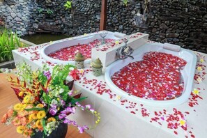 Ein Wellnessbereich mit zwei großen Badewannen, gefüllt mit Wasser und Rosenblüten, umgeben von einer Steinmauer und einem kleinen Teich.