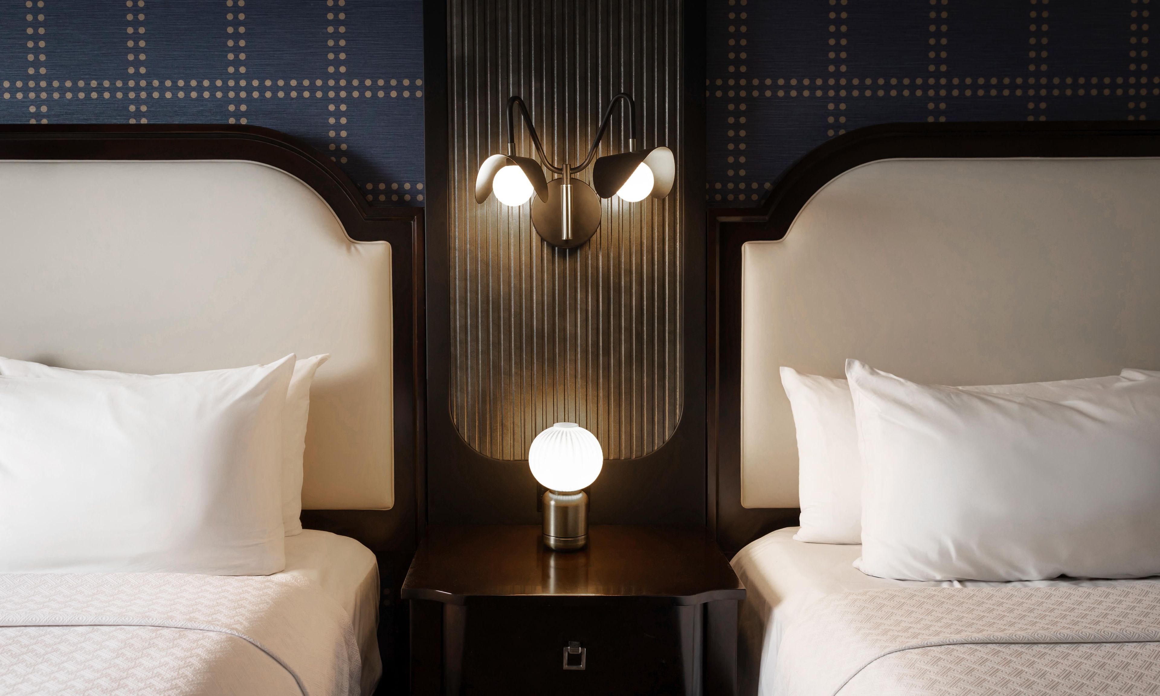 A hotel room with two beds, a nightstand with a lamp, and a wall with vertical lines.