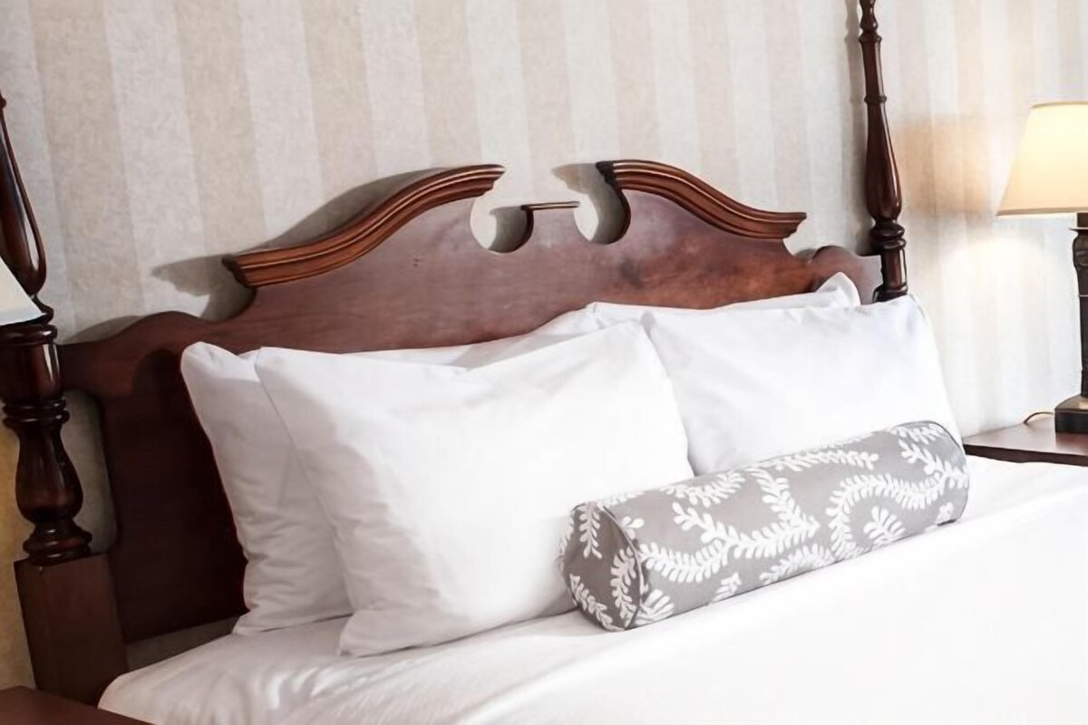Una cama con cabecera de madera, almohadas blancas y un cojín estampado.