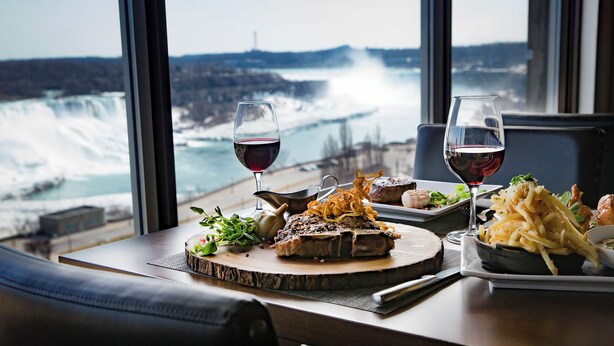 A table with a meal and two glasses of red wine, overlooking a coastal view with waves crashing against rocks. - The Brock Niagara Falls Fallsview, Tapestry by Hilton (Niagara Falls)