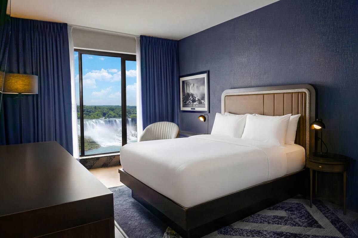 Habitación de hotel con una cama grande, un escritorio, una silla, una mesita de noche con lámpara, un cuadro enmarcado en la pared y vista a una cascada por la ventana.