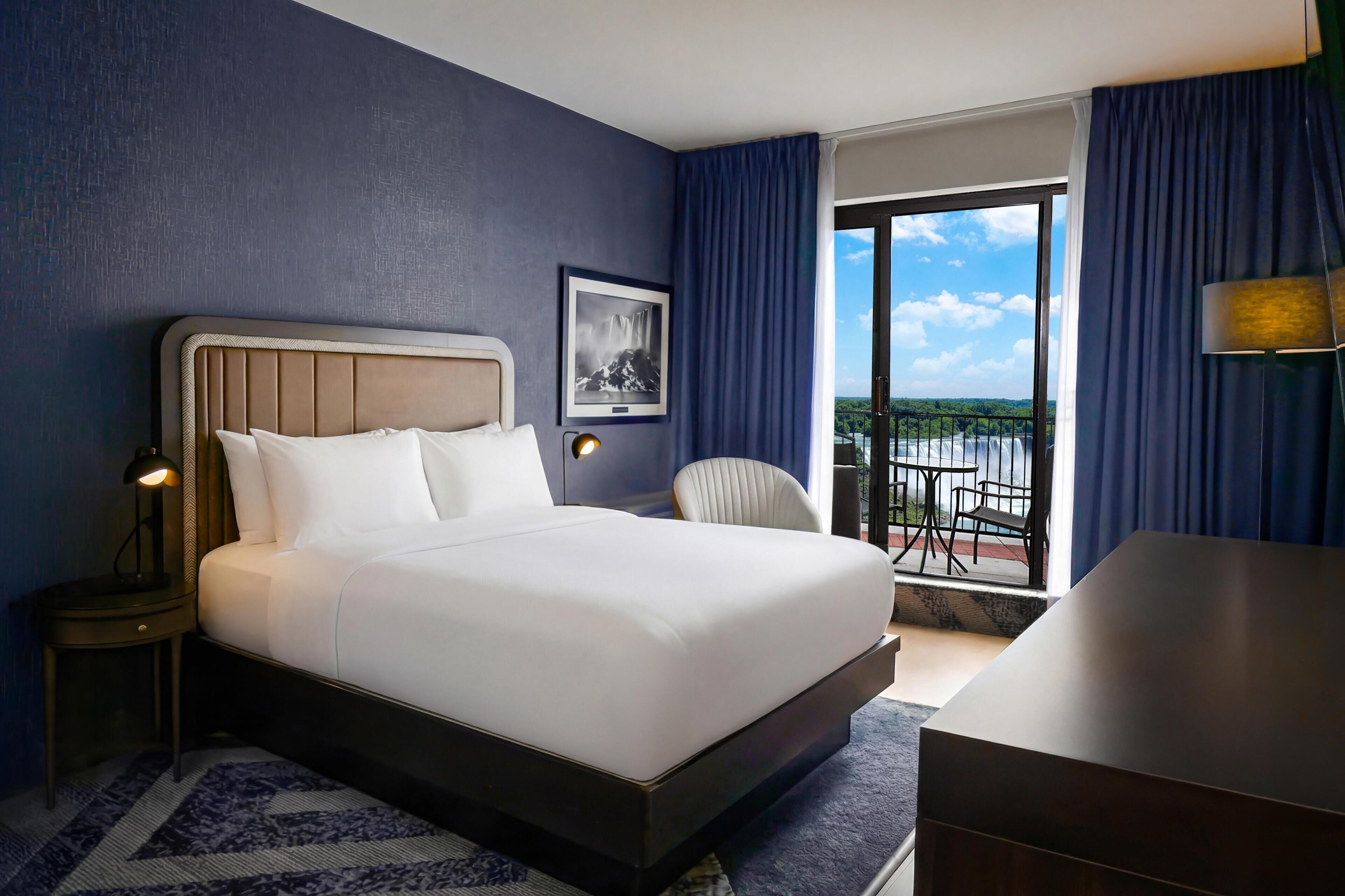 Quarto de hotel com cama, escrivaninha, cadeira, mesa de cabeceira, luminária, quadro e vista para o exterior.