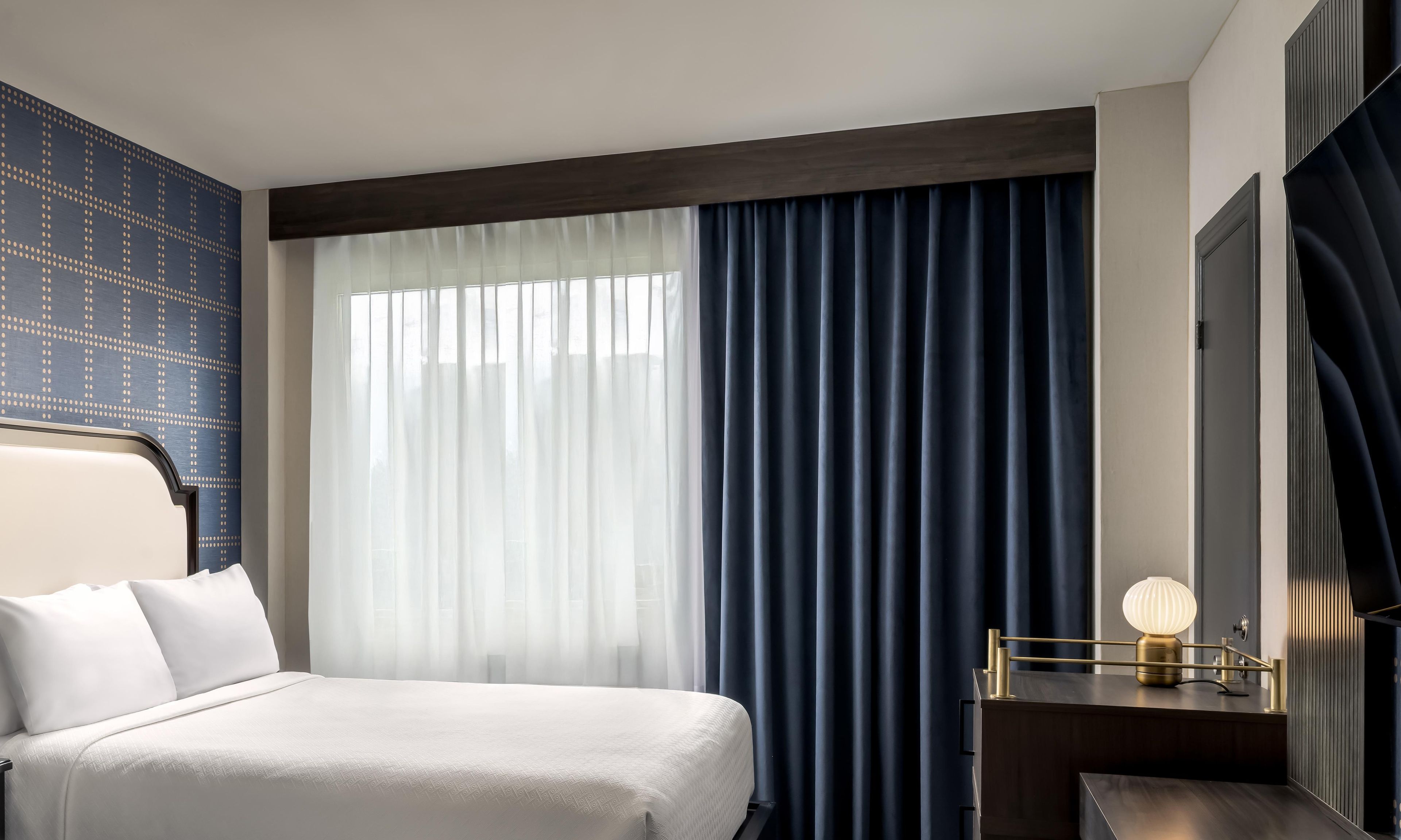 Une chambre d’hôtel avec un lit, une table de chevet avec une lampe, une fenêtre avec des rideaux et un mur orné d’un motif géométrique.