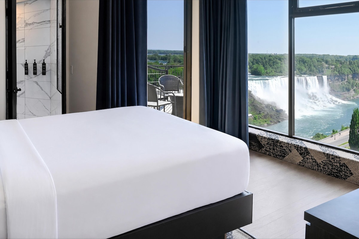 Habitación de hotel con una cama grande, vistas a una cascada y un balcón con mobiliario exterior.