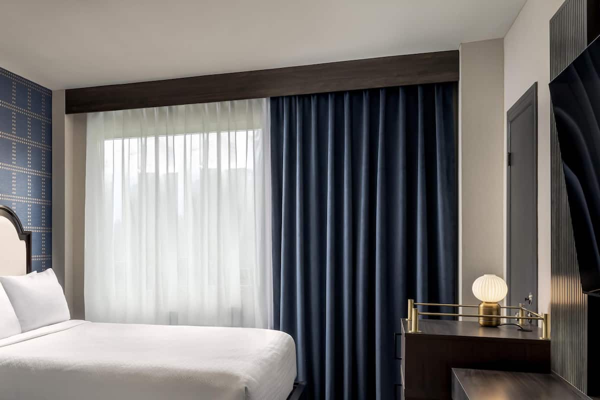 Una habitación de hotel con una cama, un escritorio con lámpara, un ventanal con cortinas y una pared con un diseño geométrico.