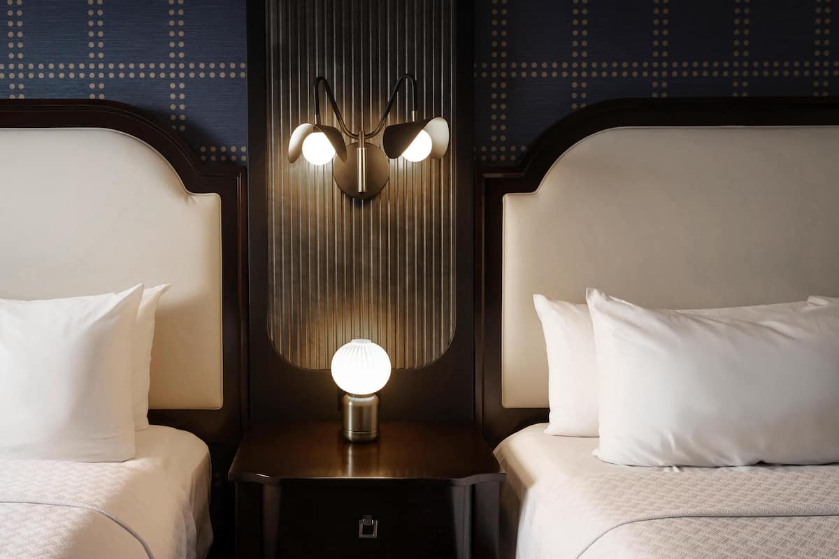 Habitación de hotel con dos camas, cada una con un almohadón blanco y una mesita de noche con una lámpara.