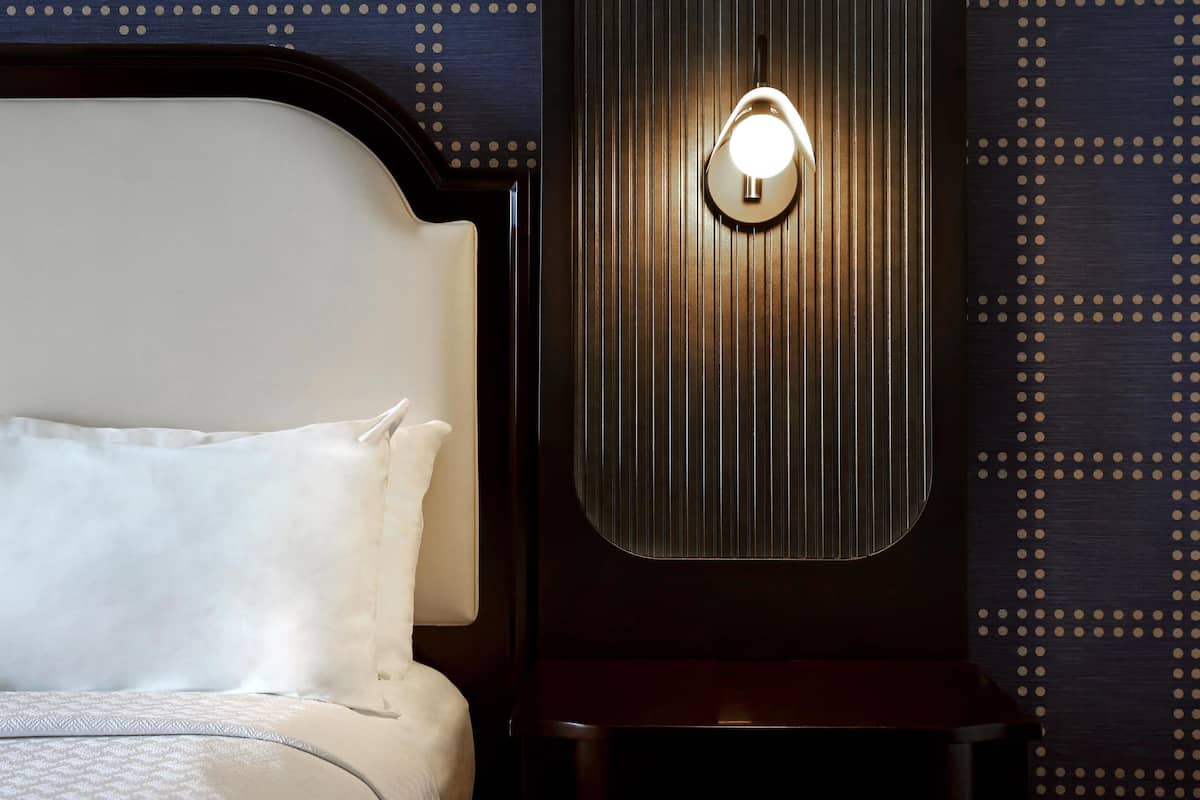 Una cama bien hecha con sábanas blancas, cabecera, mesita de noche y lámpara de pared.