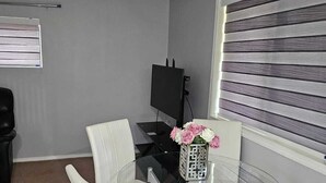 A modern room with a glass table and four white chairs, a decorative vase with flowers, a flat-screen TV, and a window with blinds.