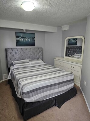 Een slaapkamer met een gestoffeerd hoofdeinde, een gestreept sprei, een dressoir met lades en een spiegel.